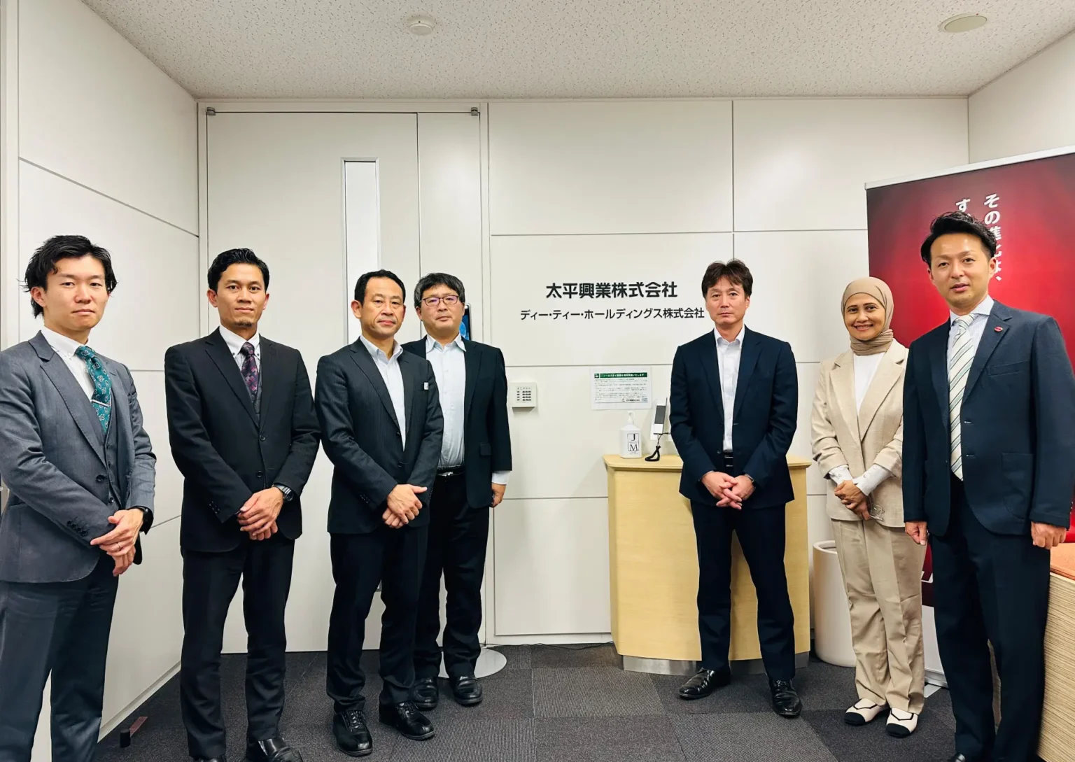 A group of six professionally dressed individuals pose together in an office setting with a wooden reception desk.