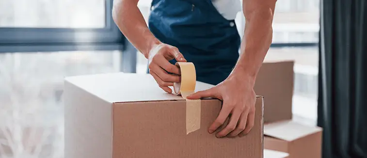 A person in blue overalls uses tape to seal a cardboard box.