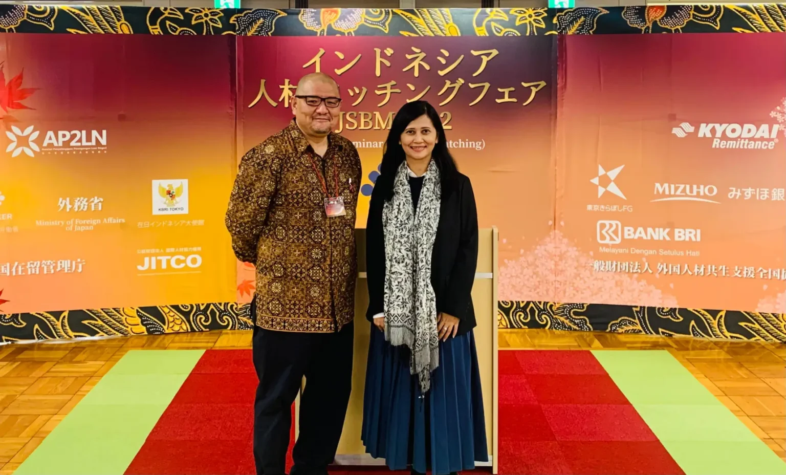 Two individuals stand in front of a colorful backdrop featuring logos and text in Japanese related to a human resource matching event.