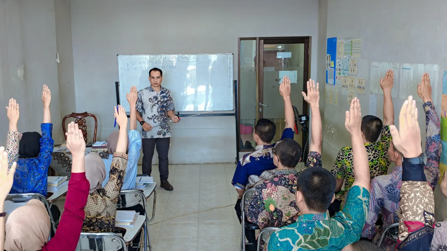 A classroom scene where students with raised hands engage with a teacher presenting in front of a whiteboard.